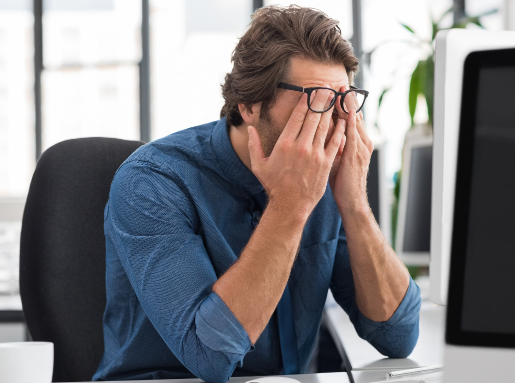 Ein gestresster Mann im blauen Hemd sitzt am Schreibtisch im Büro und hält sich erschöpft die Augen unter seiner Brille.