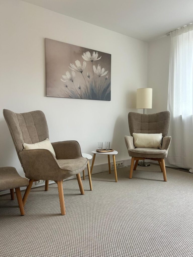 Hell eingerichteter Coaching-Raum von Resilienz Coach Sabine Maier in München Maxvorstadt mit zwei modernen beigefarbenen Sesseln, einem Beistelltisch und einem beruhigenden Wandbild mit weißen Blumen auf grauem Teppichboden.