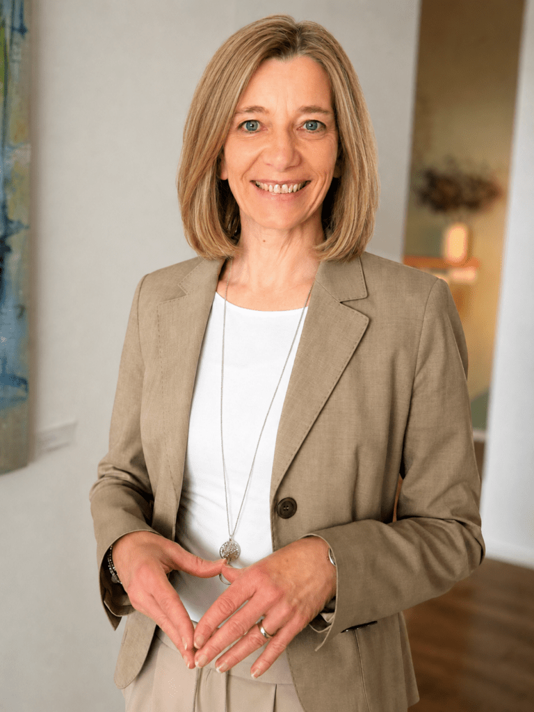 Portrait of a smiling woman in a beige blazer and white top, standing in a bright hallway.